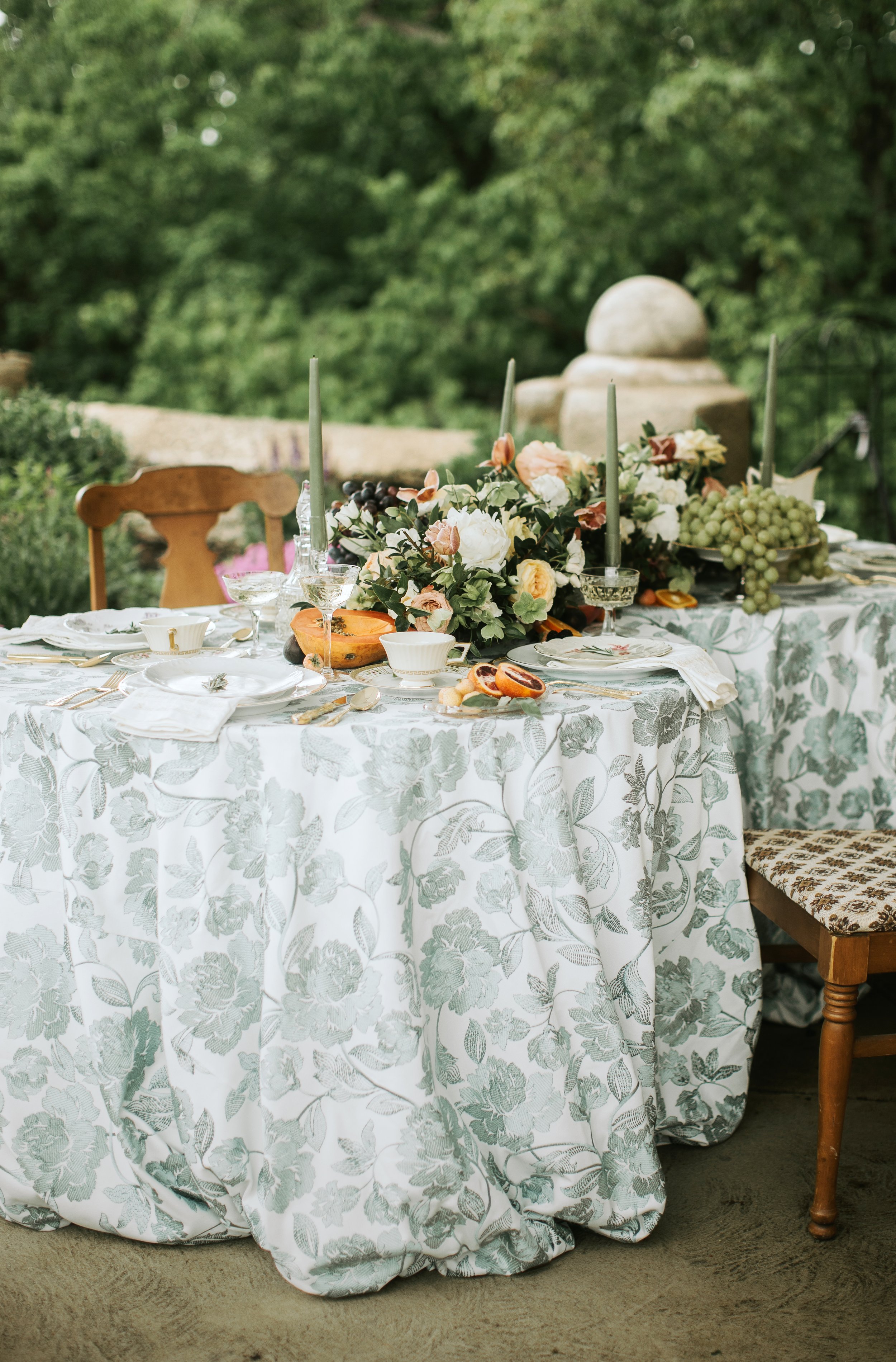 A round outdoor table with a white floral tablecloth, decorated with a floral centerpiece, candles, and various dishes and glasses, set for a meal in a garden setting.